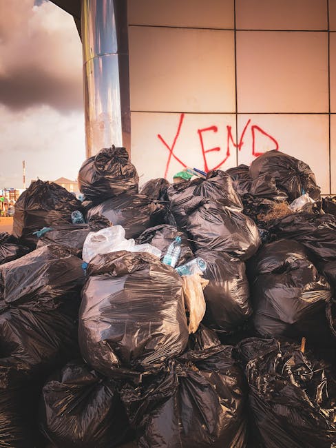 A large pile of black rubbish bags filled with waste, stacked against a modern building with reflective tiles on the exterior wall, which has the word 'XEND' spray-painted in red graffiti. Several dis