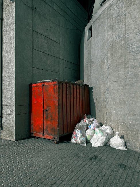 A row of five large, rectangular wheelie bins is positioned along a sidewalk adjacent to a brick wall. The bins are made of durable plastic with textured, ribbed lids and body surfaces, each painted i