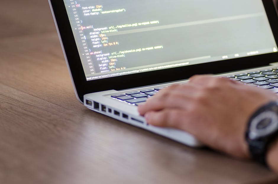 Close-up view of a modern laptop placed on a surface with a dark background, displaying a code editor with multiple lines of programming code on the screen. The code includes syntax highlighting with