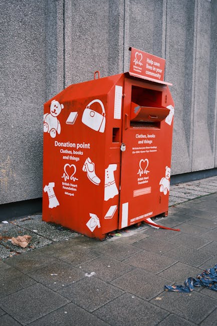 A red donation drop-off bin located on a paved pavement beside a grey concrete wall. The bin is designed with illustrations of various items such as a teddy bear, handbag, bottle, clothing, and shoes