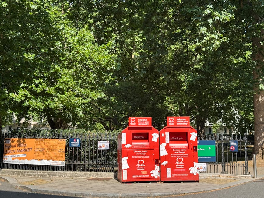 A red donation drop-off bin located on a paved pavement beside a grey concrete wall. The bin is designed with illustrations of various items such as a teddy bear, handbag, bottle, clothing, and shoes
